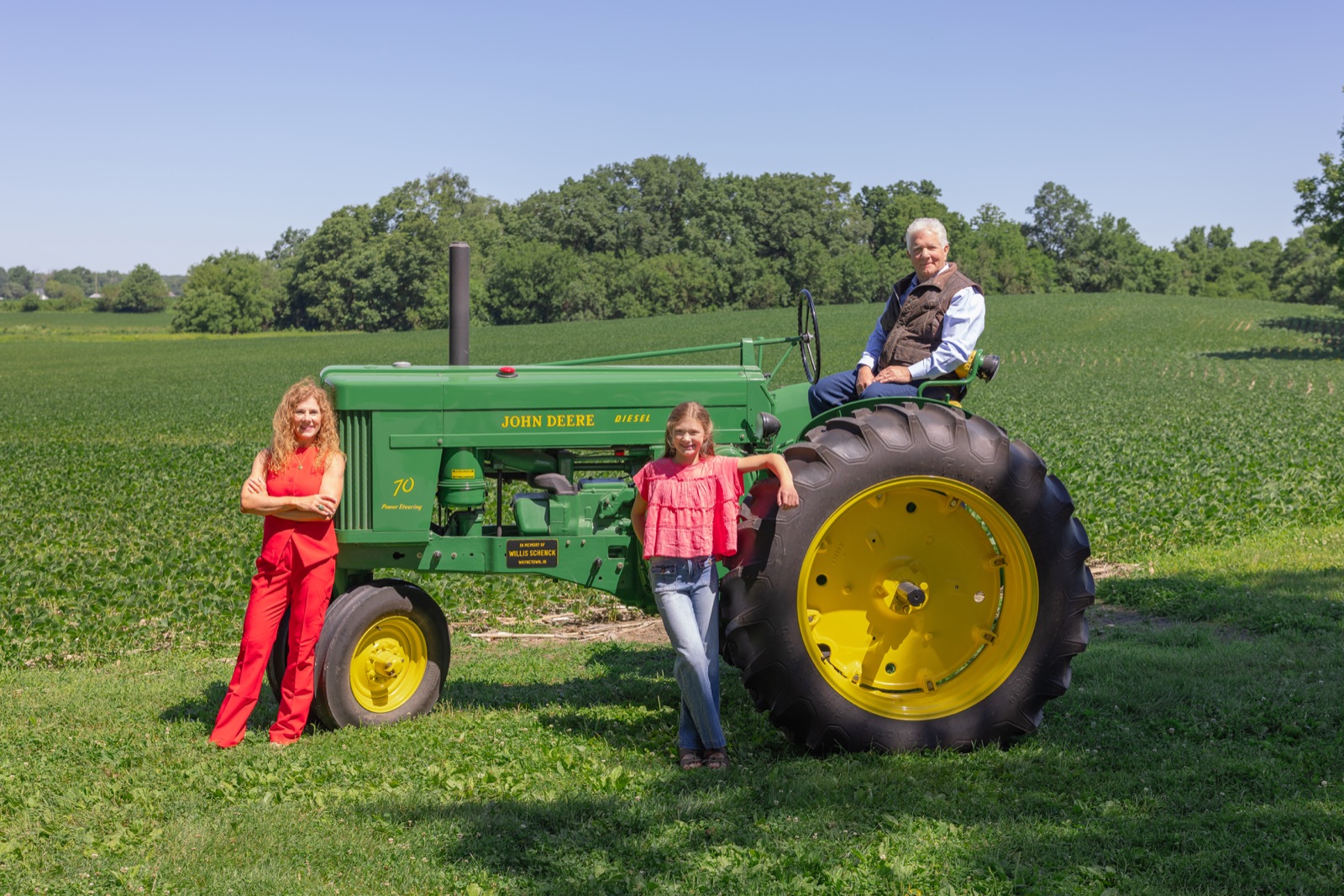 Sara, her daughter, and her father together beside a vintage John Deere tractor bearing a memorial plaque for Willis Schenck.