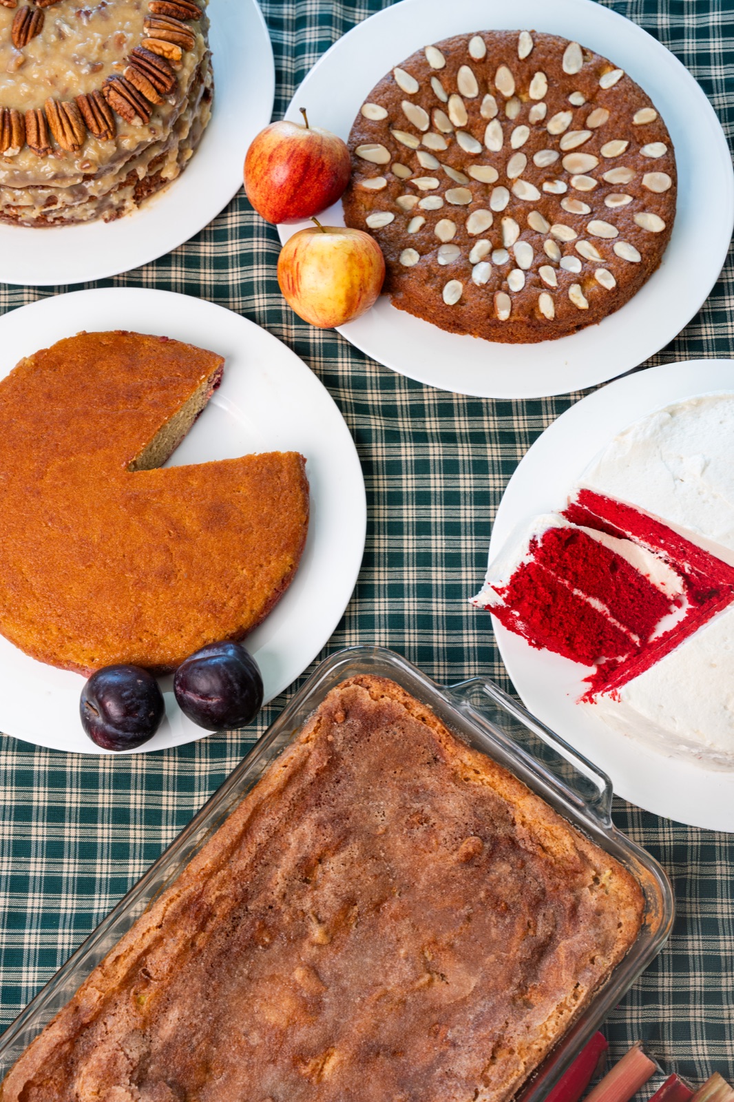 Overhead spread of cakes, apples, and plums on a green plaid tablecloth.